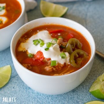 a close up of chicken soup topped with sour cream and green onion in a white bowl next to lime wedges