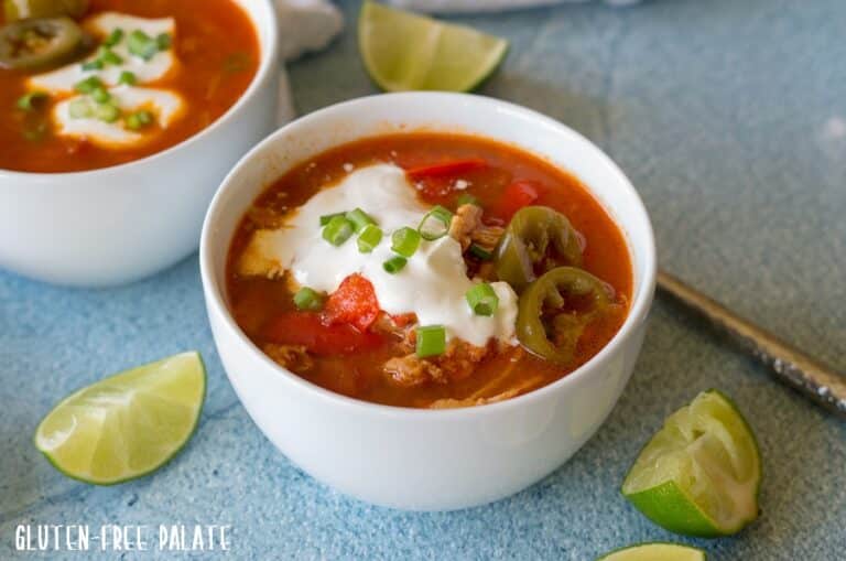 a close up of chicken soup topped with sour cream and green onion in a white bowl next to lime wedges
