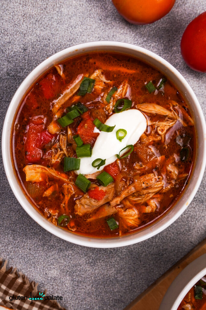 A top-down view of chicken fajita soup in a bowl, featuring shredded chicken, diced tomatoes, sour cream, and green onions, placed next to fresh produce.