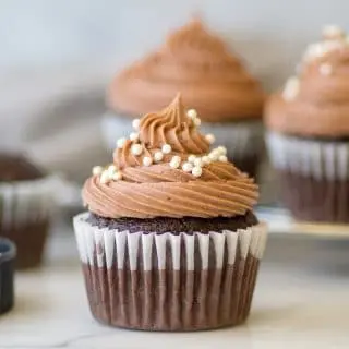 a close up of a gluten-free chocolate cupcake with chocolate frosting and white pearl sprinkles on top