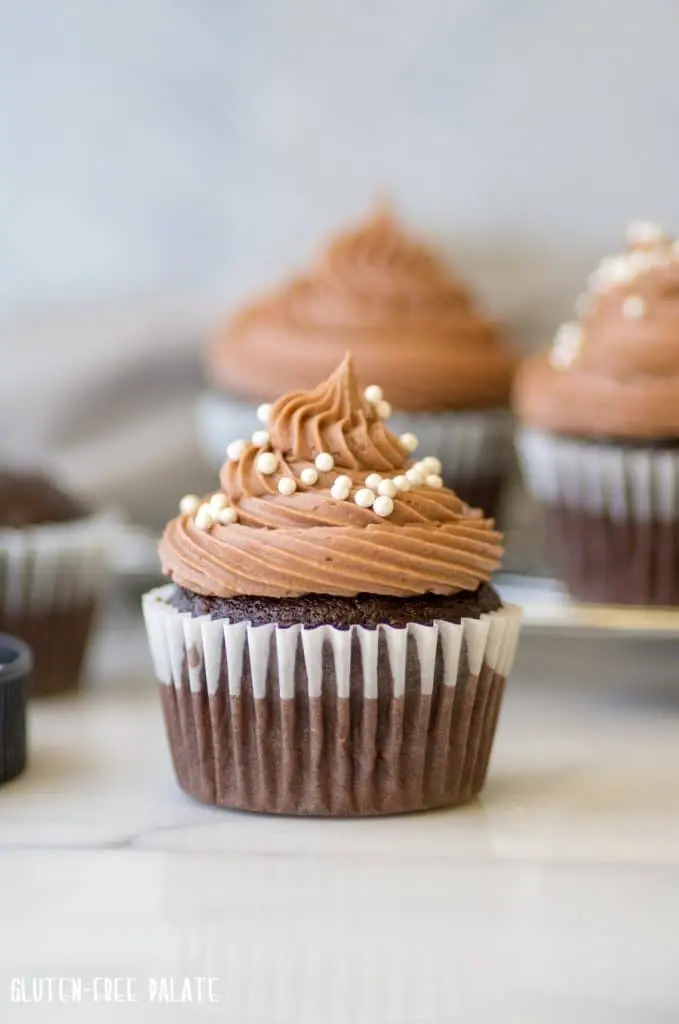 a close up of a gluten-free chocolate cupcake with chocolate frosting and white pearl sprinkles on top