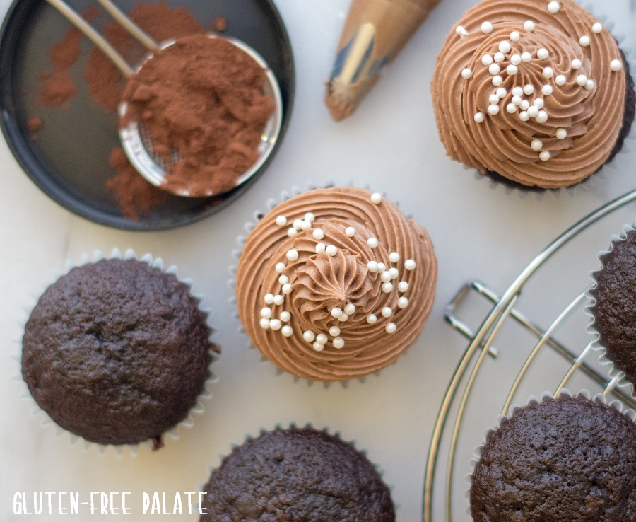 a close up of a top down view of gluten-free chocolate cupcakes with chocolate frosting and white pearl sprinkles on top nex to unsweetened cocoa on a sifter
