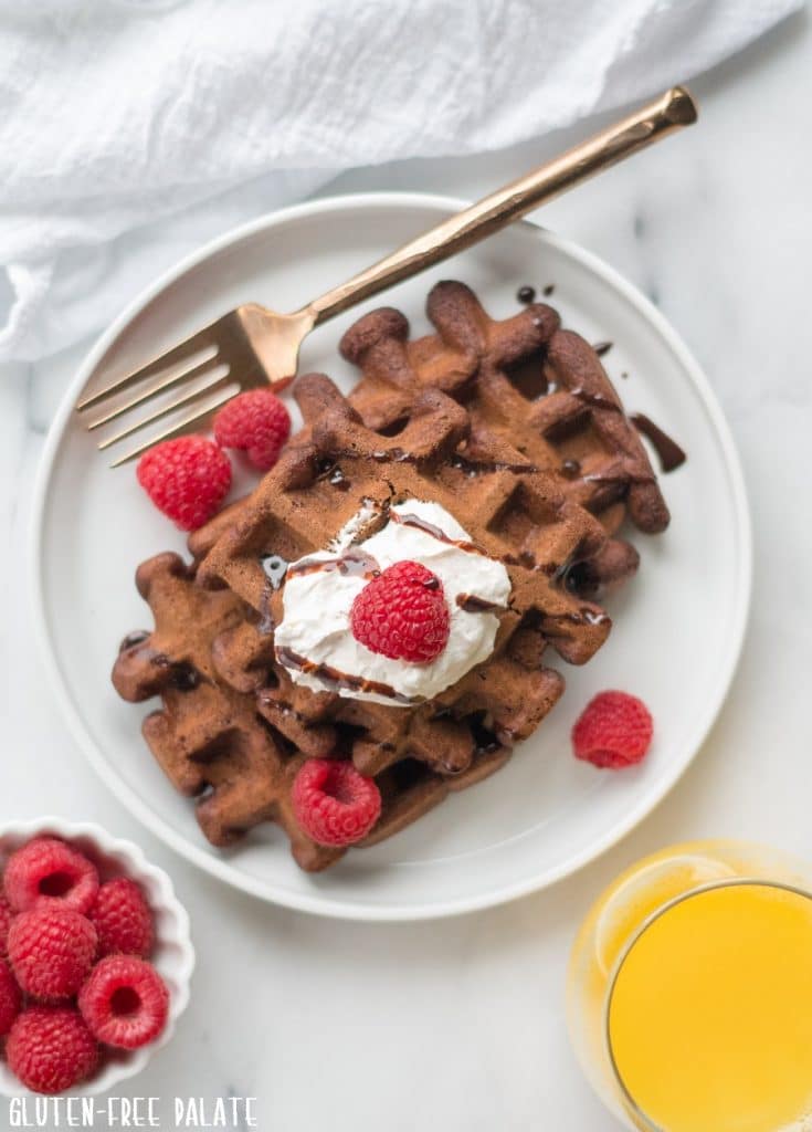 top down view of chocolate waffles on a white plate with raspberries next to a bowl of raspberries and a glass of orange juice