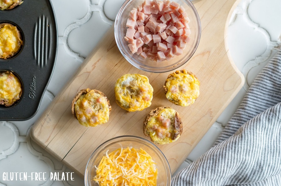 Mini Quiches on a cutting board next to a bowl of cheese and a bowl of chopped ham