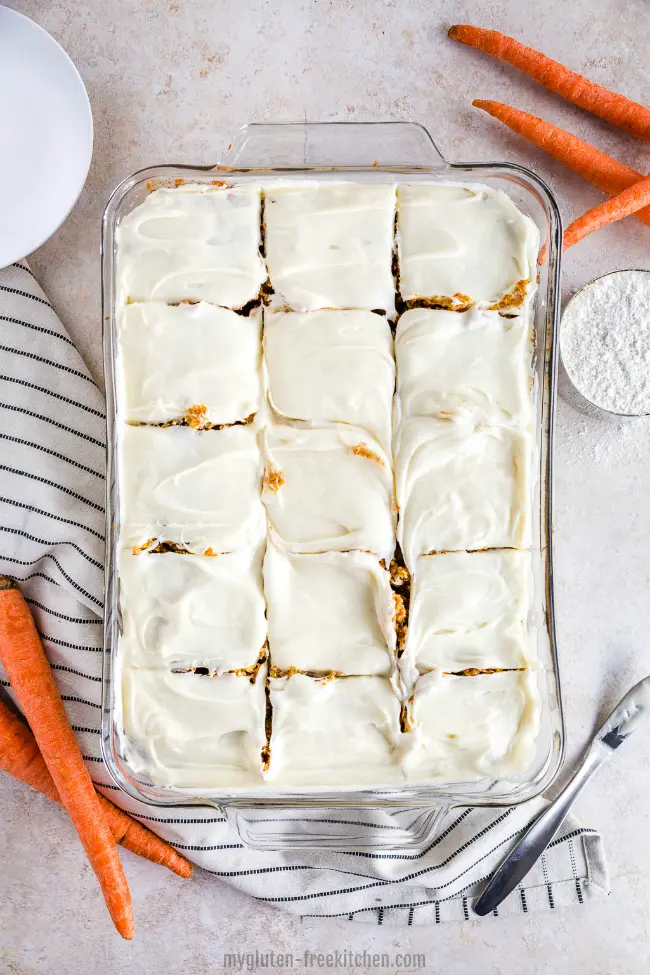 A glass pan with gluten-free carrot cake and white frosting, with carrots and a stripe napkin next to it