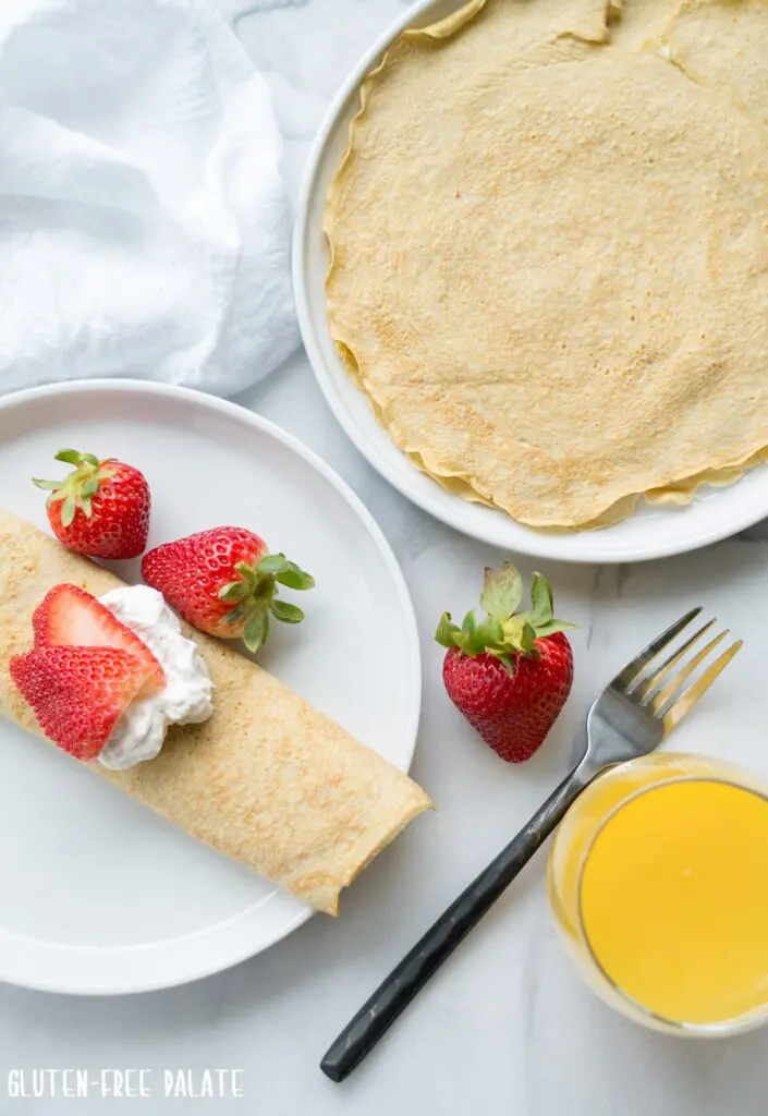 a crepe on a white plate, topped with whipped cream and strawberries next to a crepe and a glass of orange juice