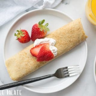 a crepe on a white plate, topped with whipped cream and strawberries next to a crepe and a glass of orange juice