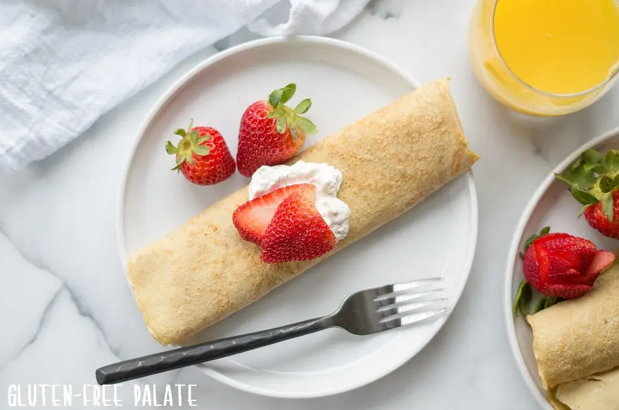 a crepe on a white plate, topped with whipped cream and strawberries, with a fork next to a glass of orange juice