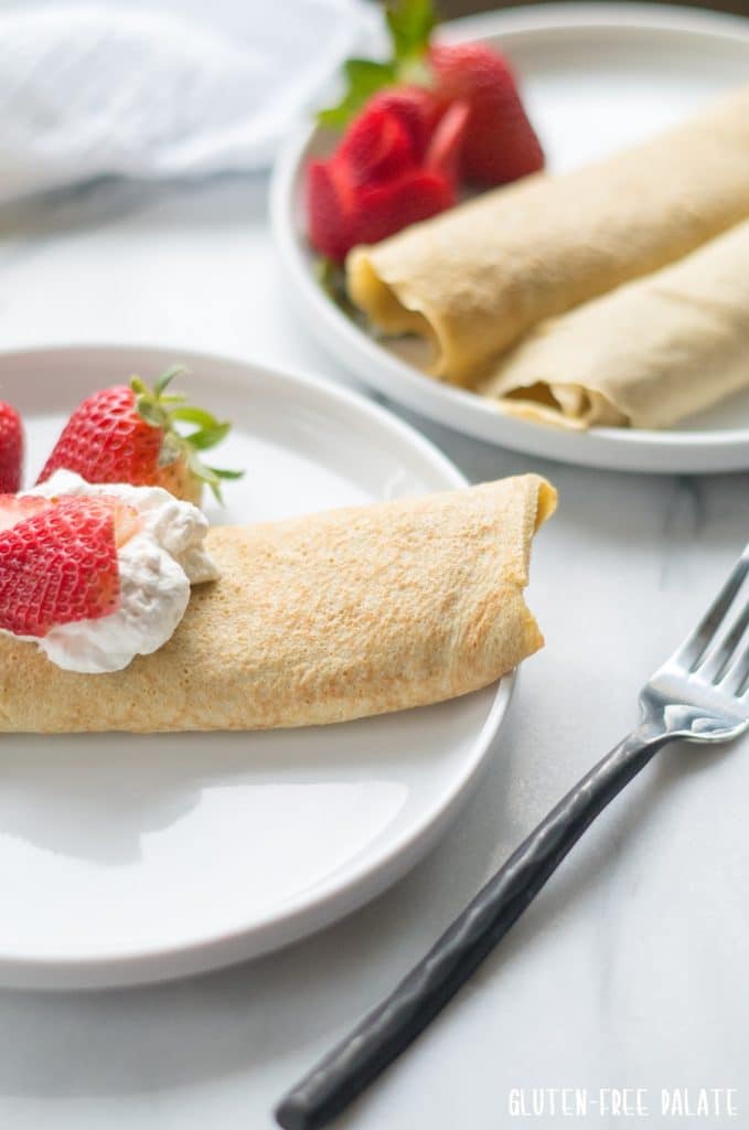 side view of a crepe on a white plate, topped with whipped cream and strawberries next to a fork