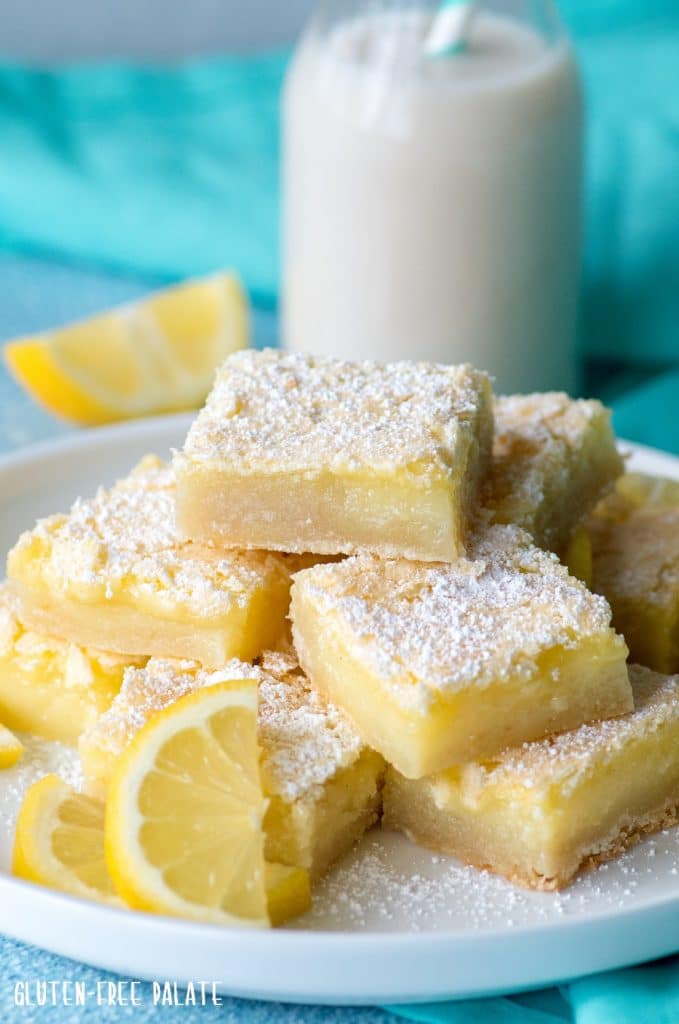 gluten free lemon bars stacked on a white plate with a jar of milk, a slice of lemon, and a blue napkin in the background