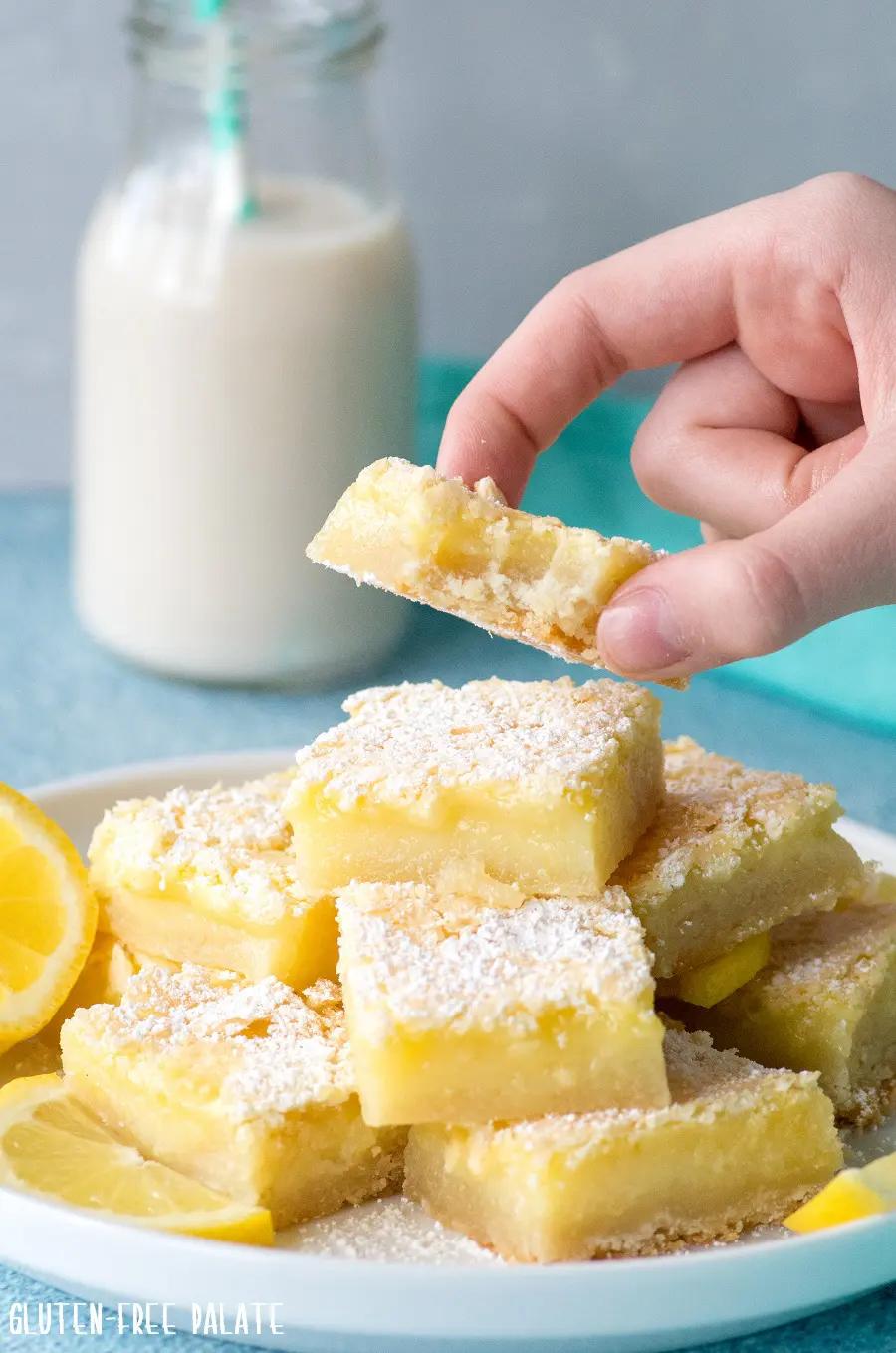 a hand holding a gluten-free lemon bars with a bite out, over a plate of lemon bars