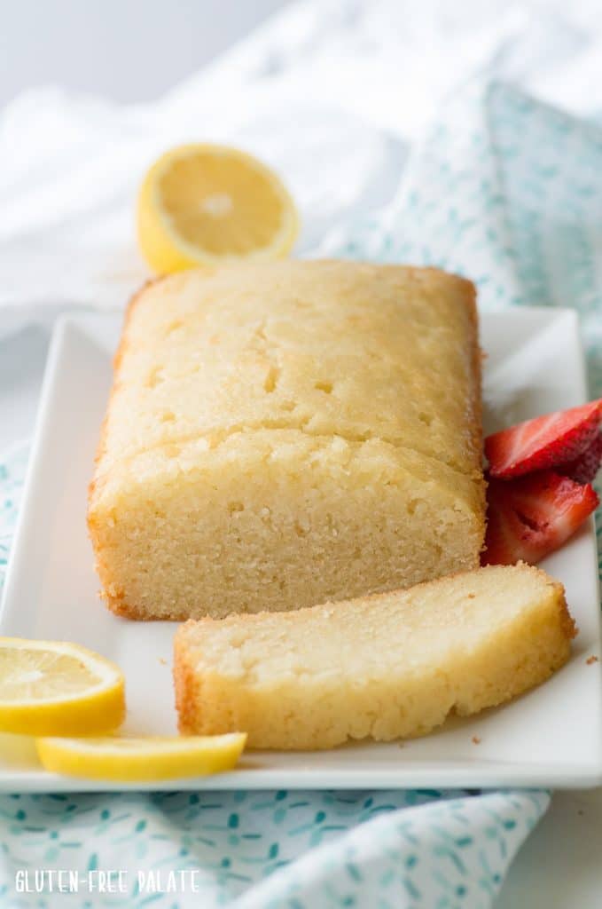 a side view of a Gluten-Free Lemon Bread loaf with a couple of lemon bread slices, on a white plate with lemon and strawberry slices on the side