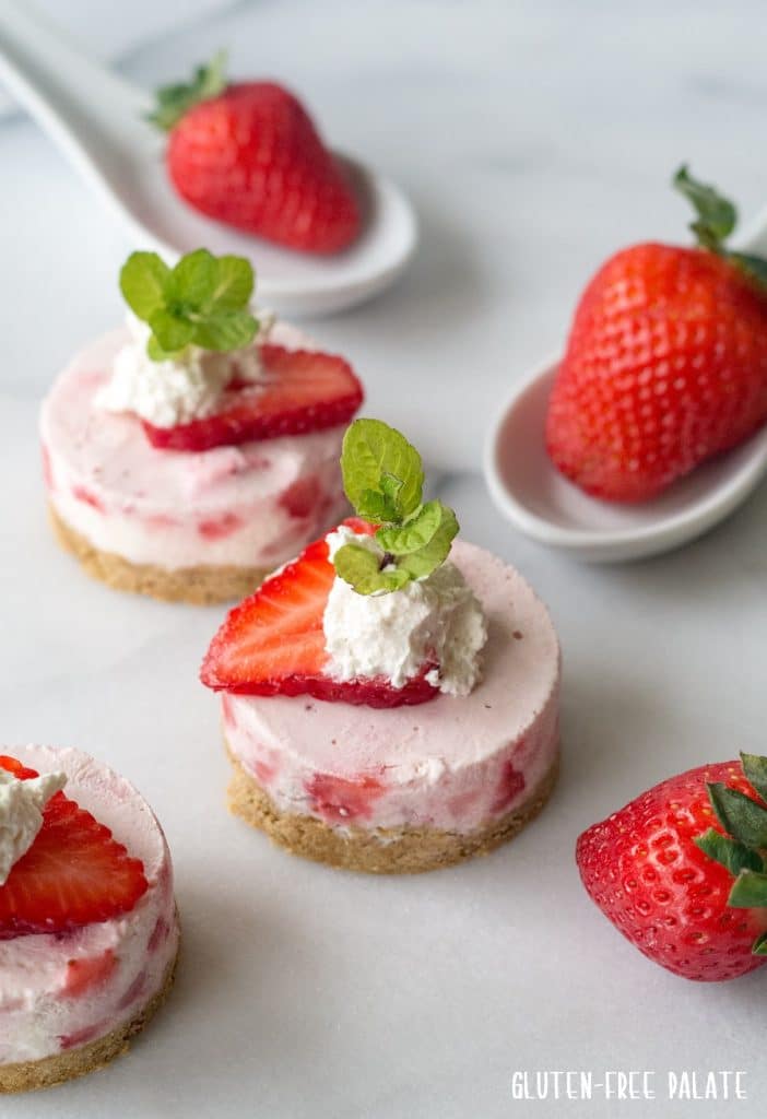 side view of mini strawberry cheesecakes with whipped cream and a mint leaf on top, next to strawberries