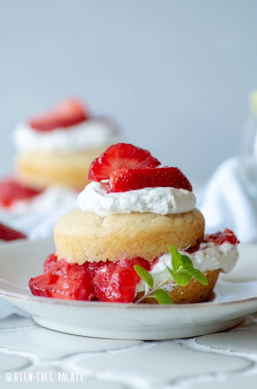 a close up of a Gluten-Free Strawberry Shortcake with whipped cream and sliced strawberries on a white plate with a spring on mint on the side