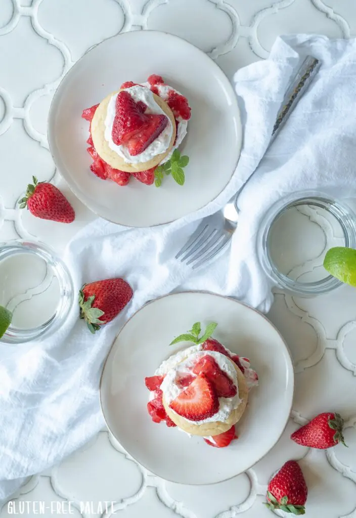 top down view of two a Gluten-Free Strawberry Shortcakes with whipped cream and sliced strawberries on white plates with a spring on mint on the side