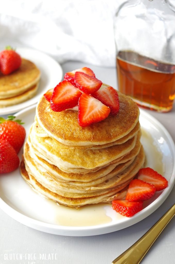 Gluten-Free Pancakes stacked on a white plate, topped with strawberries next to a jar of maple syrup.