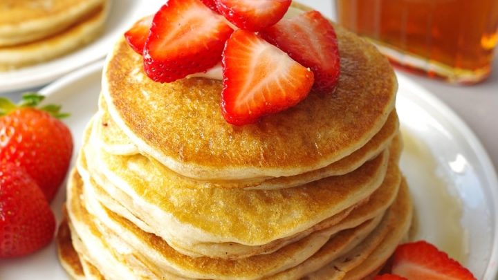 a close up of Gluten Free Pancakes stacked on a white plate, topped with strawberries next to a jar of maple syrup