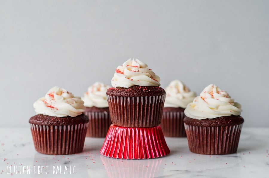 gluten free red velvet cupcakes topped with white frosting and red sugar, one is sitting on a shiny red paper liner