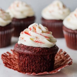 a close up of a gluten free red velvet cupcake topped with white frosting and red sugar, with the paper liner peeled down