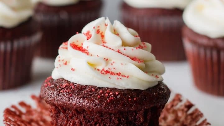 a close up of a gluten free red velvet cupcake topped with white frosting and red sugar, with the paper liner peeled down