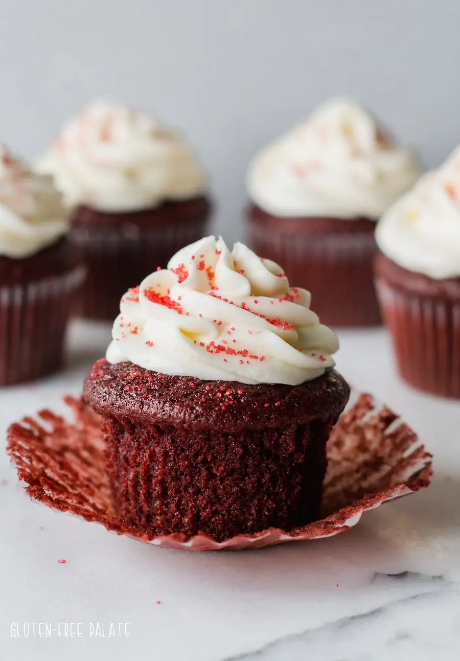 a gluten free red velvet cupcake topped with white frosting and red sugar, with the paper liner peeled down