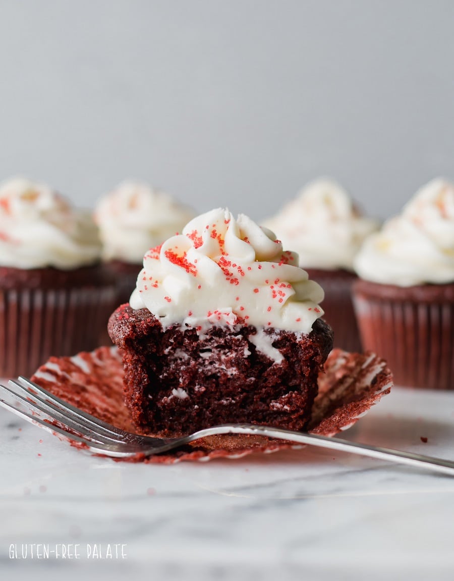 red velvet cupcake topped with white frosting and red sugar, with a bite taken out and a fork