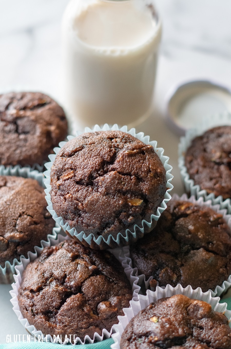 close up of Grain-Free Chocolate Zucchini Muffins on a white background with a blue kitchen towl and a jar of milk