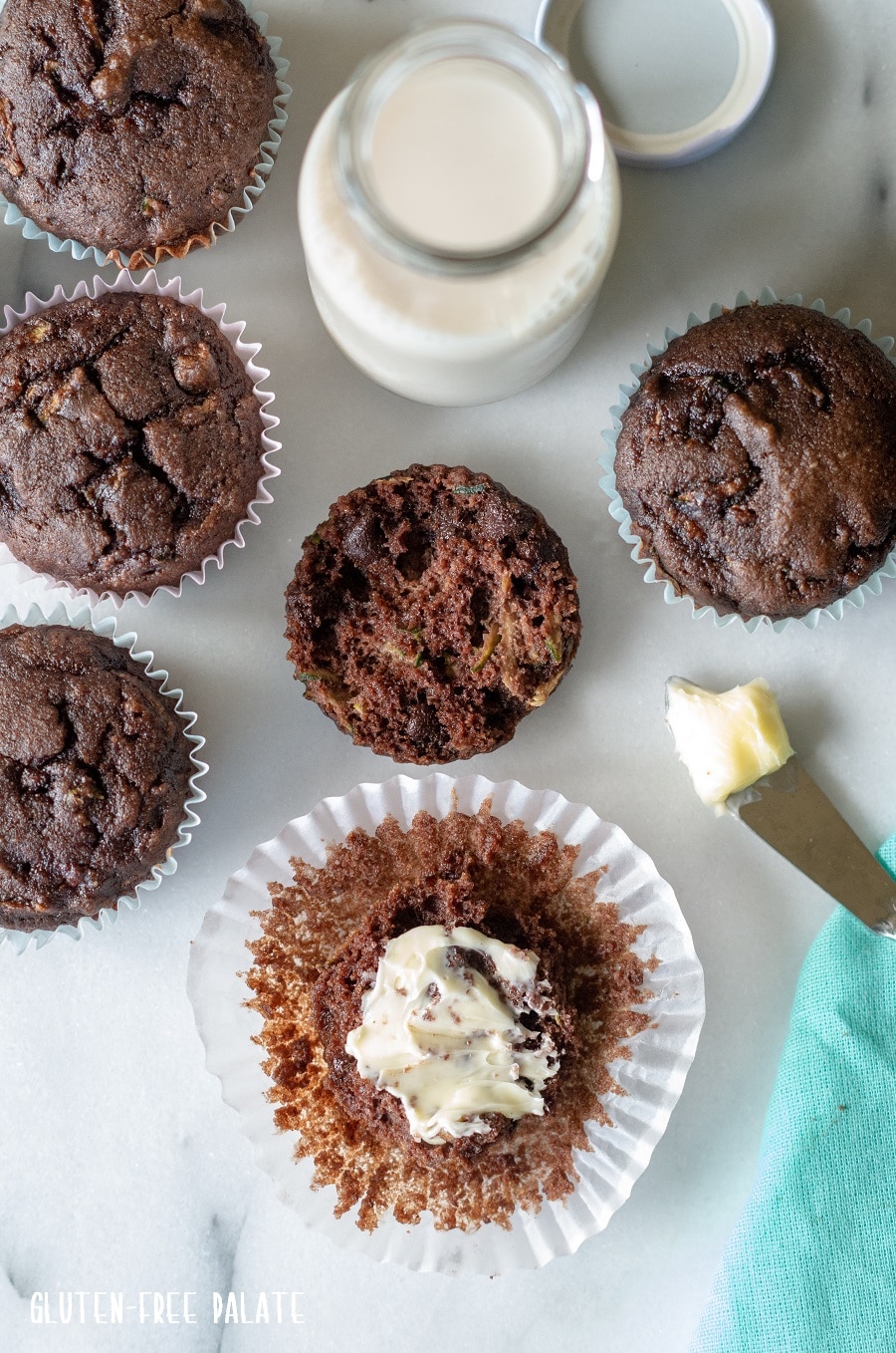 top down view of Grain-Free Chocolate Zucchini Muffins on a white background with a blue kitchen towl and a jar of milk, one muffin has butter spread on top