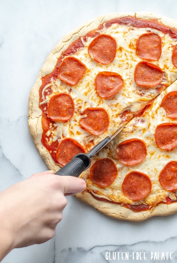 a hand cutting a slice of Gluten Free Pizza topped with cheese and pepperoni