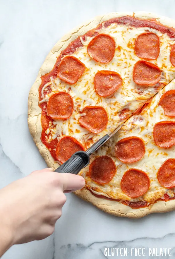 a hand cutting a slice of Gluten-Free Pizza topped with cheese and pepperoni