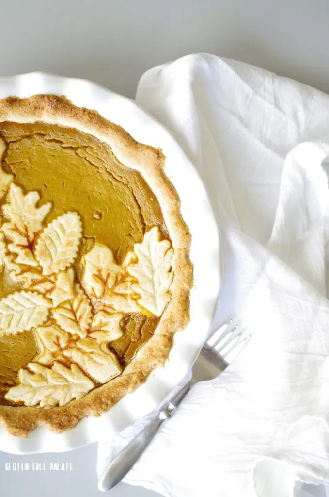 a close up of gluten free pumpkin pie in a white pie pan with white pumpkins and a white napkin