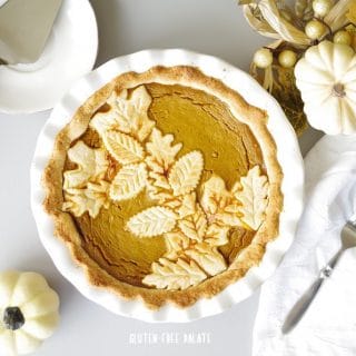 a close up of gluten-free pumpkin pie in a white pie pan with white pumpkins and a white napkin