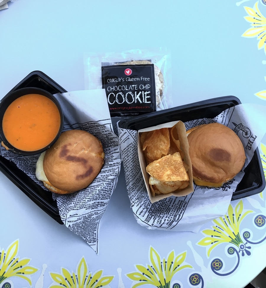 two trays with a grilled cheese sandwich on round shaped bread next to a bowl of tomato soup, and a gluten free chocolate chip cookie in a package
