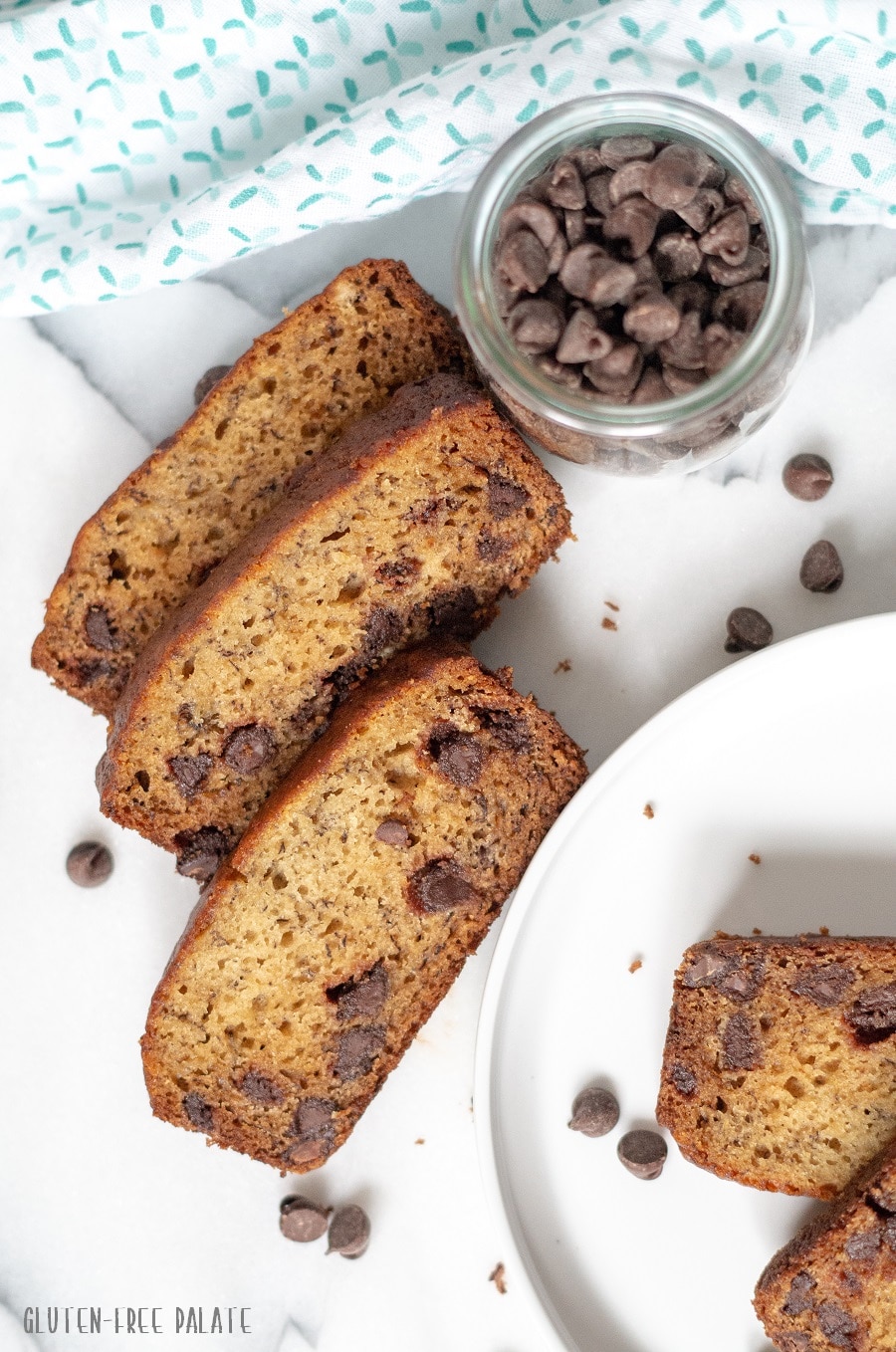 slices of banana bread next to a white plate with slices of banana bread, and a jar of chocolate chips next to a blue and white towel