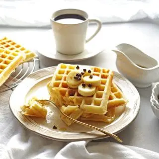 close up of three waffles on a cream colored plate with a bite taken out on a fork, a cup of coffee in the background