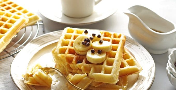 close up of three waffles on a cream colored plate with a bite taken out on a fork, a cup of coffee in the background