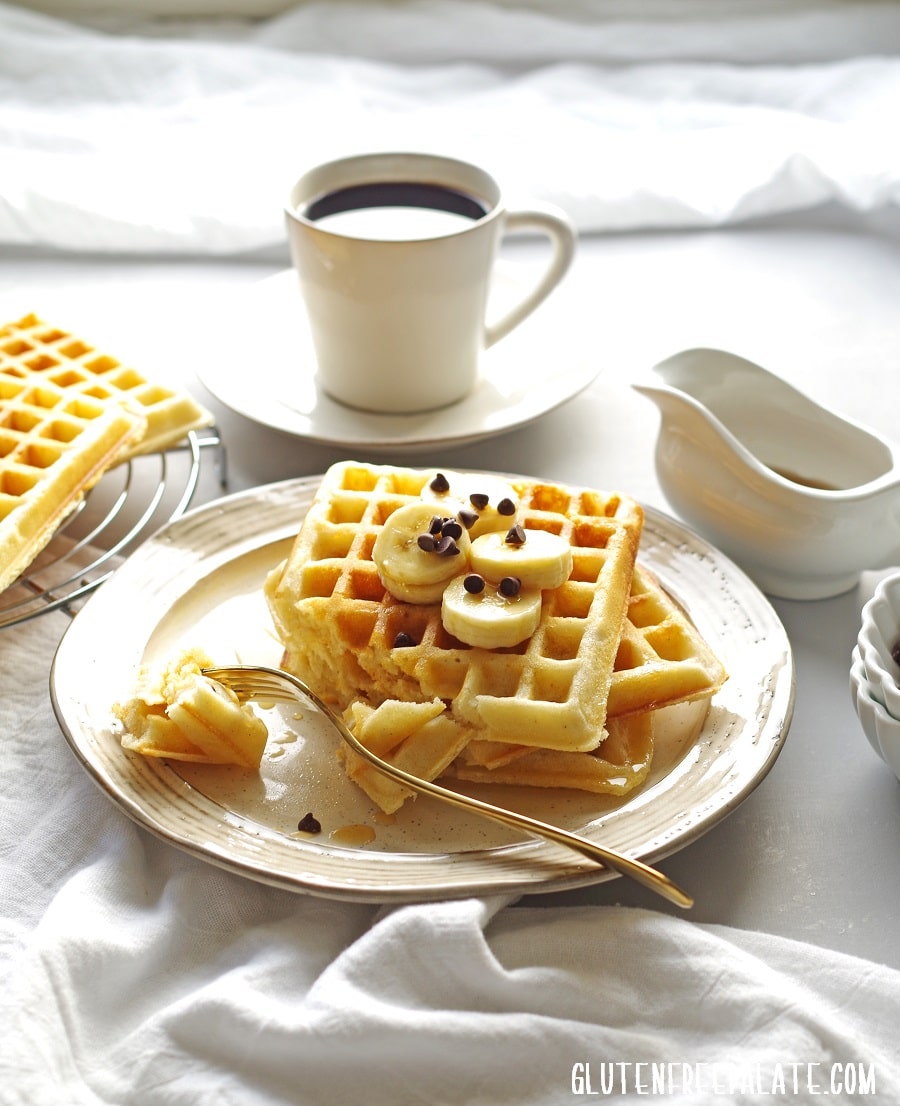 three waffles on a cream colored plate with a bite taken out on a fork, a cup of coffee in the background