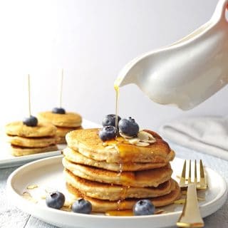 close up of pancakes stacked on a white plate, topped with blueberries and maple syrup being poured on top