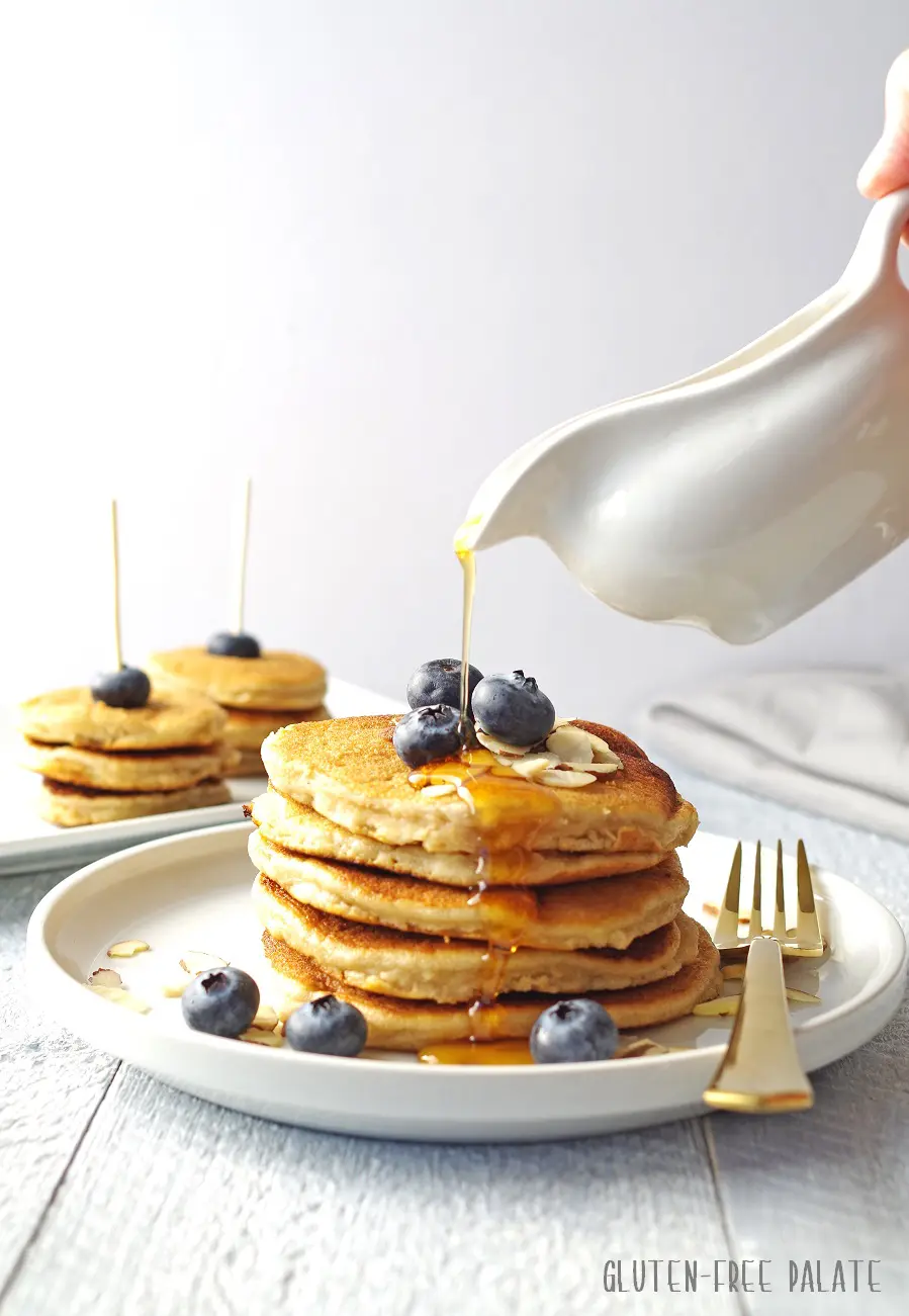 pancakes stacked on a white plate, topped with blueberries and maple syrup being poured on top