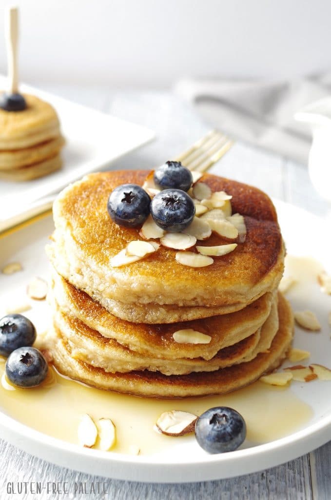 Paleo pancakes stacked on a white plate, topped with blueberries and maple syrup with sliced almonds.