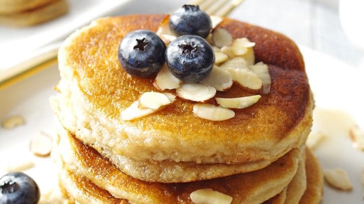 close up of pancakes stacked on a white plate, topped with blueberries and maple syrup with sliced almonds