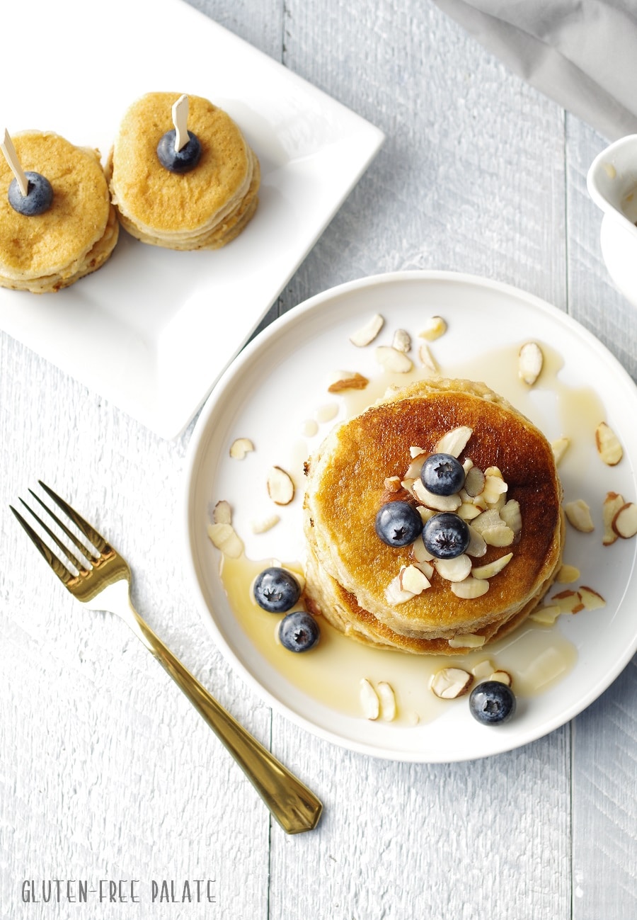 top down view of pancakes stacked on a white plate, topped with blueberries, sliced almonds and maple syrup