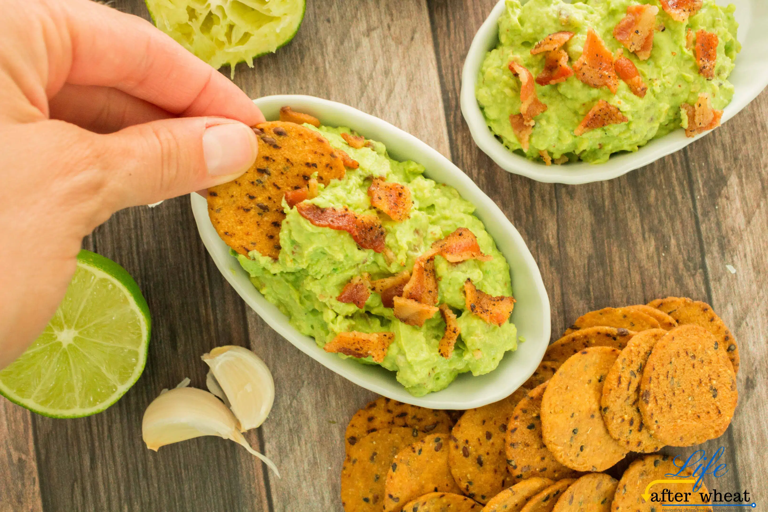 a hand dipping a chip into Roasted Avocado & Bacon Guacamole, next to lime wedges and chips