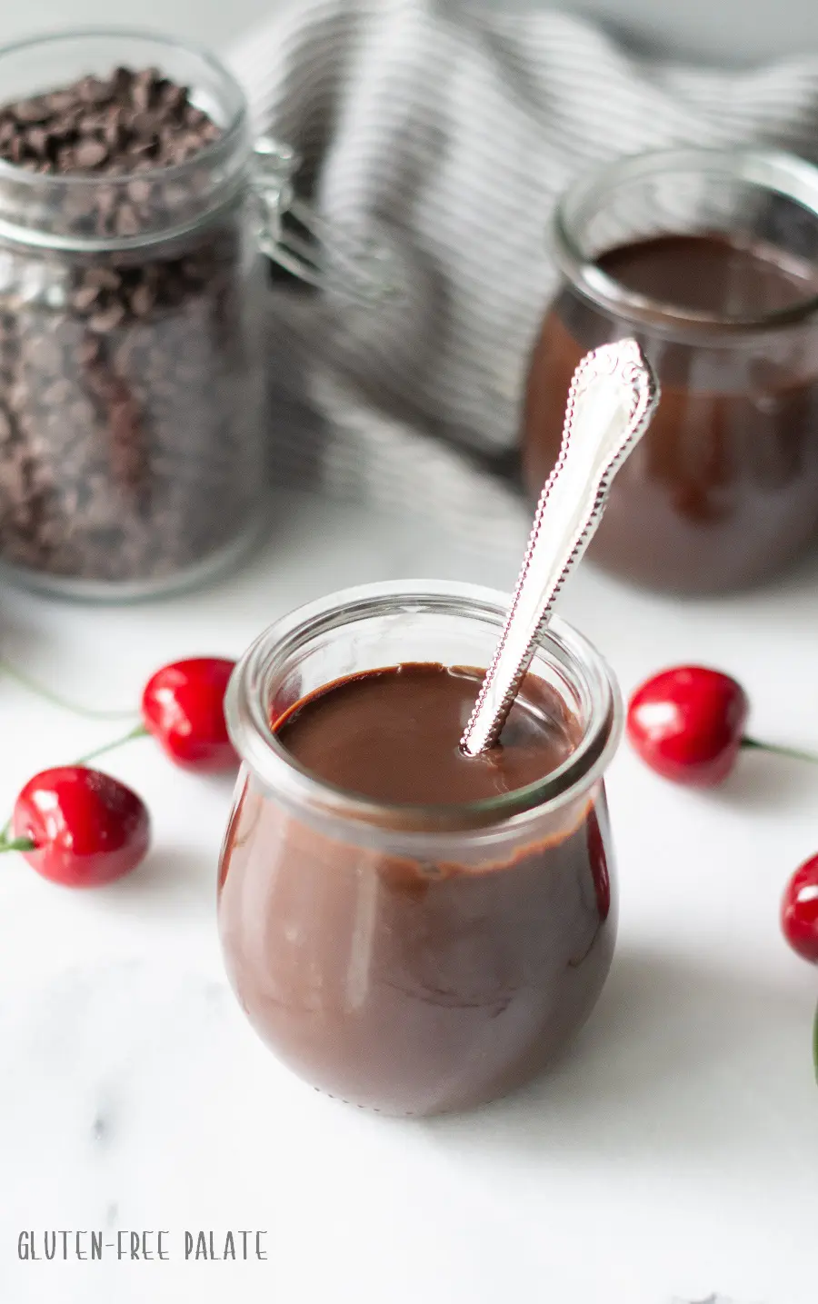 chocolate sauce in a clear jar with a spoon with cherries and a seperate jar of chocolate chips