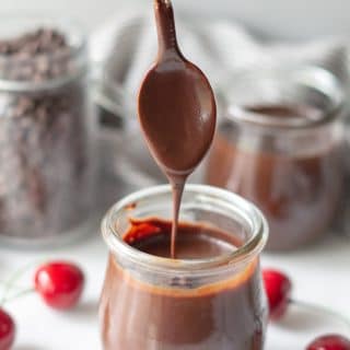 chocolate sauce in a clear jar with a spoon with cherries and a seperate jar of chocolate chips