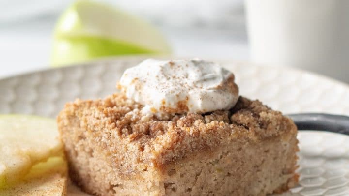 a slice of paleo apple cake with whipped cream on top, on a white plate with sliced apple