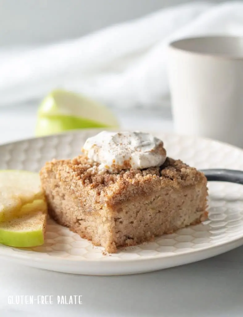 a slice of paleo apple cake with whipped cream on top, on a white plate with sliced apple