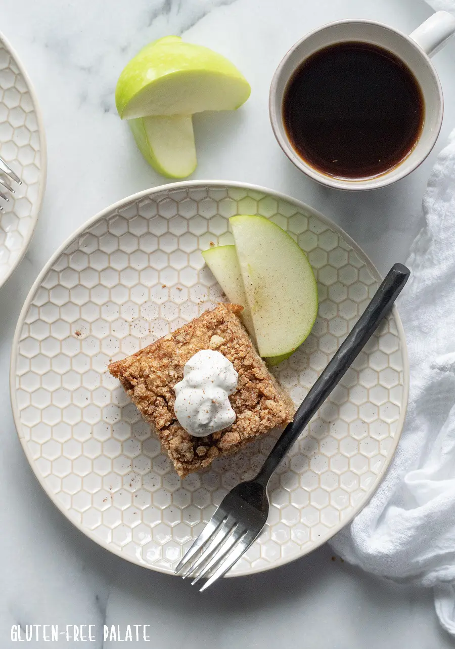 downward view of a slice of paleo apple cake with whipped cream on top, on a white plate with sliced apple, and a cup of coffee
