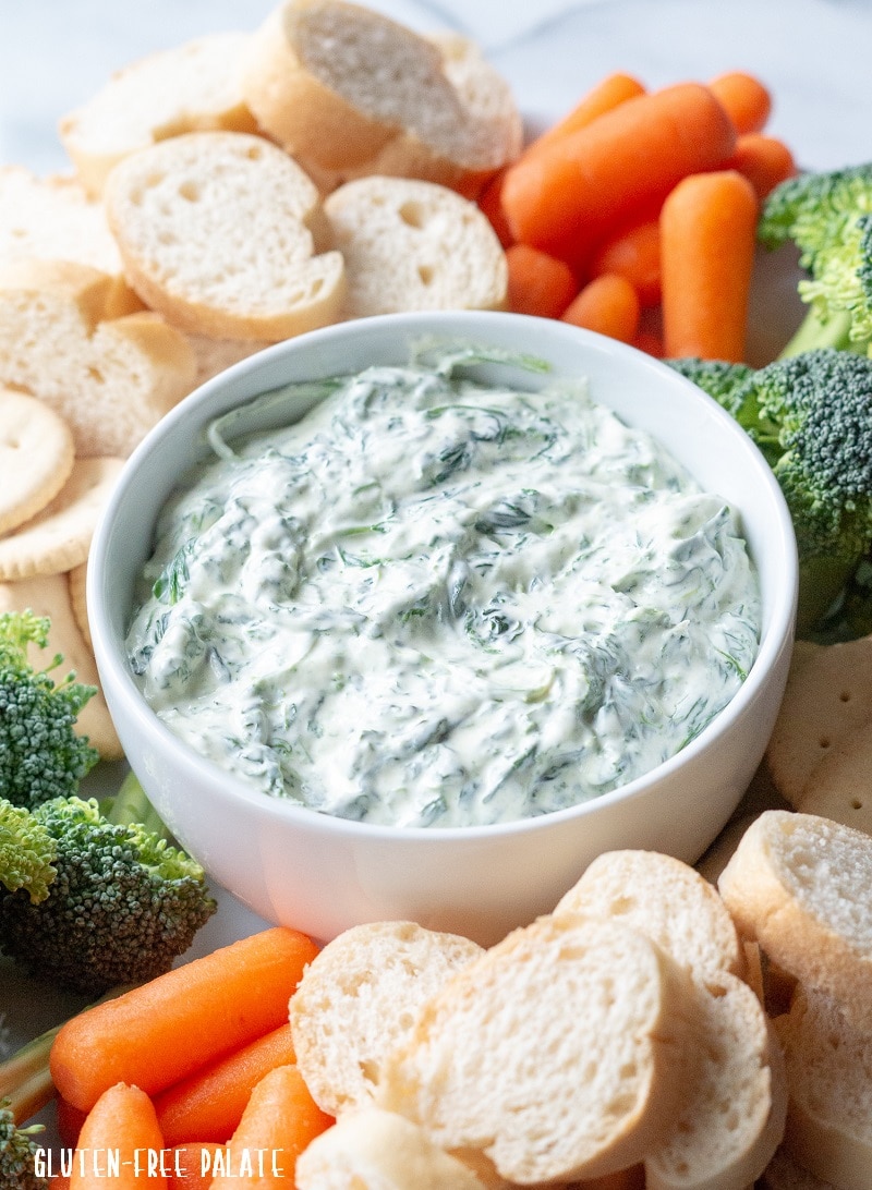 close up of a white bowl filled with spinach dip next to slices of bread, carrots, and broccoli