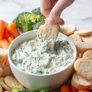 a hand dipping a slice of bread into a bowl of spinach dip, next to broccoli and carrots and bread slices